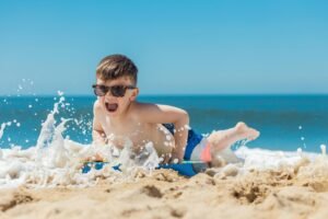A young boy in sunglasses joyfully splashes on the beach, embracing the summer waves and sun.